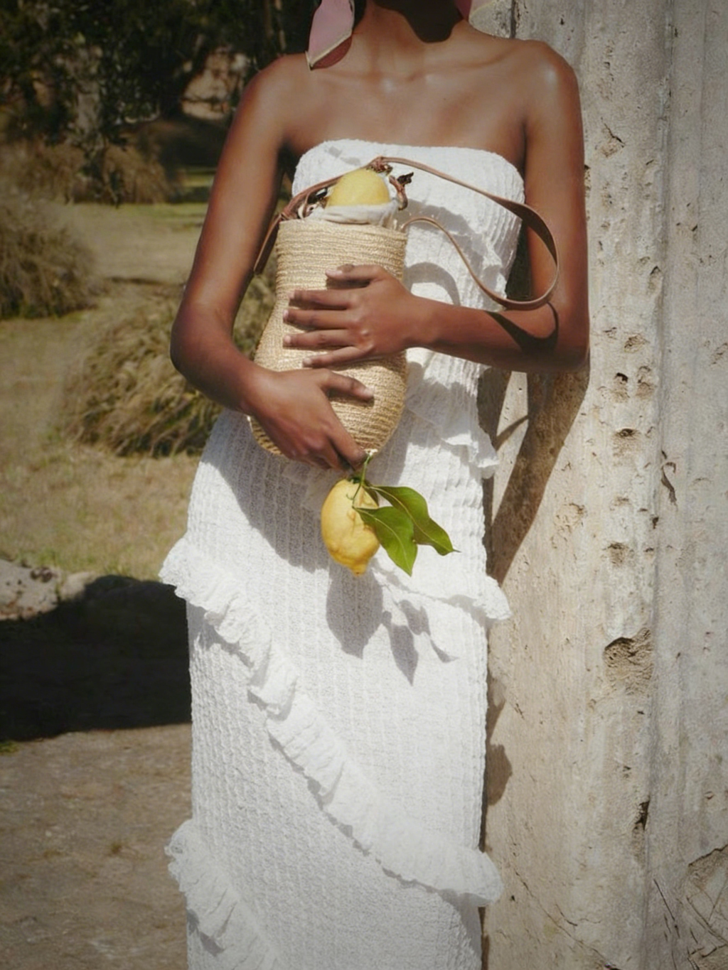 Robe Tris Elyth Édition : robe longue bustier blanche en maille gaufrée avec volants diagonaux, portée par une femme tenant un panier en osier garni de citrons devant une colonne antique ensoleillée.