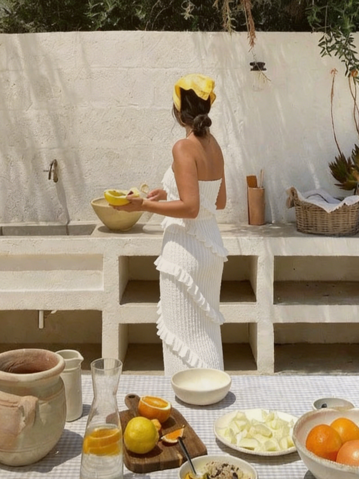Femme de dos portant la robe Tris Elyth Édition, une robe longue bustier blanche en maille gaufrée à volants, préparant des fruits dans une cuisine d'été méditerranéenne en extérieur, avec un foulard jaune dans les cheveux.
